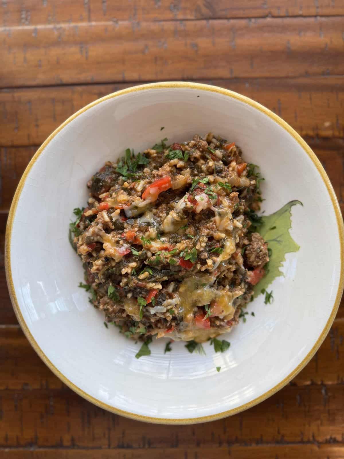 A bowl of mixed ground meat, vegetables, and melted cheese, garnished with chopped herbs on a wooden table.