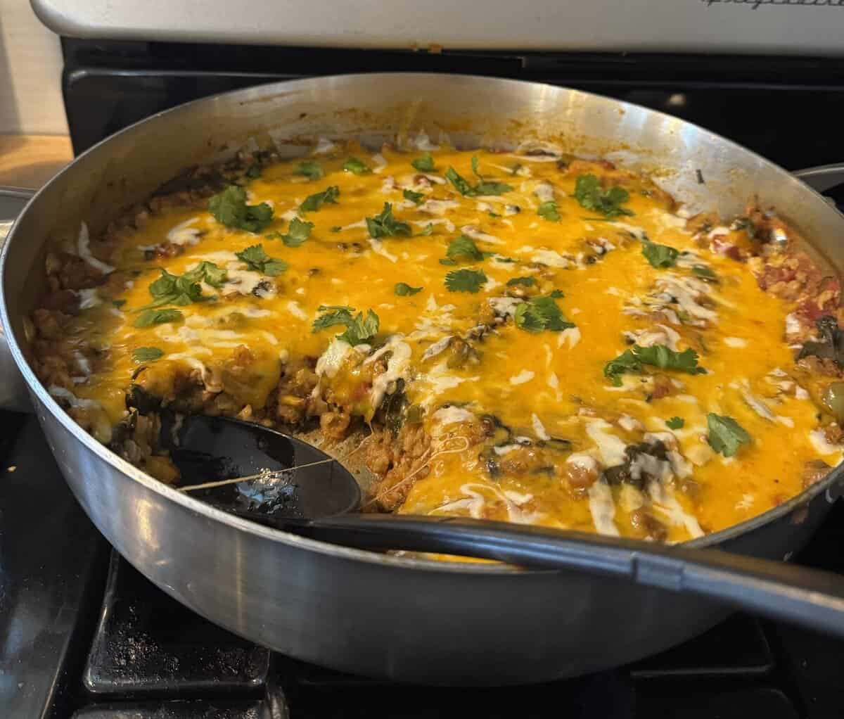 A skillet filled with cheesy casserole topped with melted cheese and fresh cilantro, with a serving spoon inside.