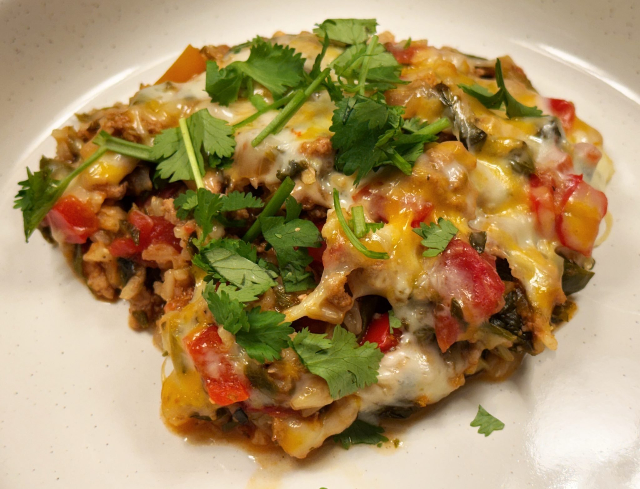 A serving of cheesy casserole with ground meat, bell peppers, and fresh cilantro on top, on a white plate.