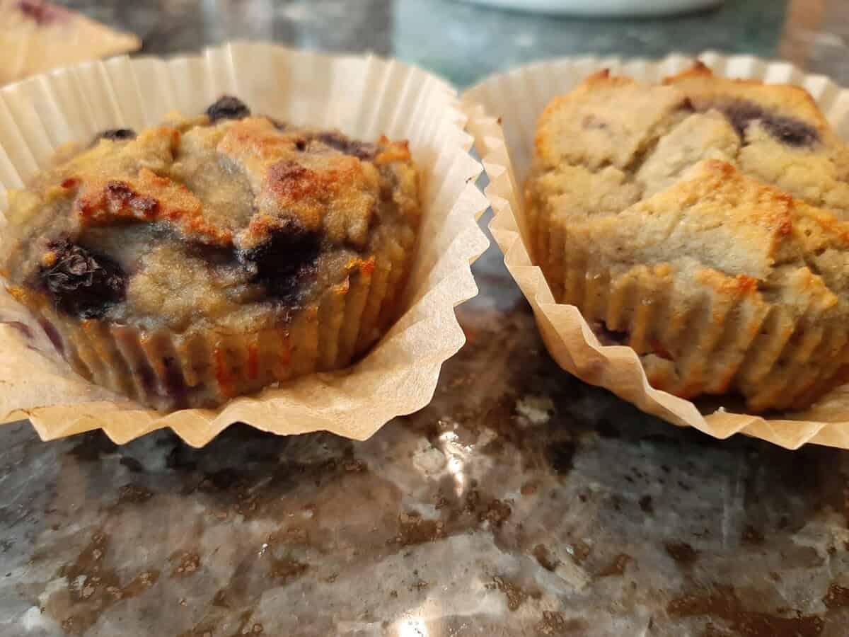 Two blueberry muffins in brown paper liners sit on a marbled countertop.