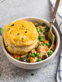 A bowl of chicken pot pie filling with peas and carrots, topped with a golden biscuit and garnished with chopped herbs.