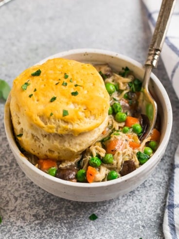 A bowl of chicken pot pie filling with peas and carrots, topped with a golden biscuit and garnished with chopped herbs.
