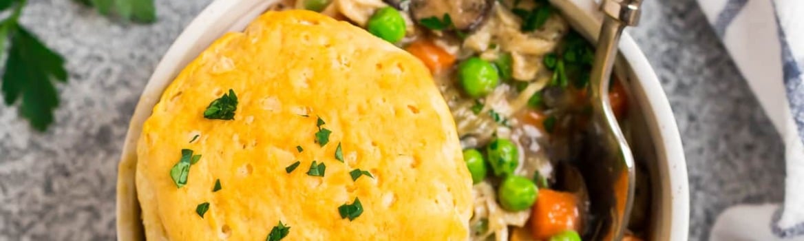 A bowl of chicken pot pie filling with peas and carrots, topped with a biscuit, and a spoon on the side.