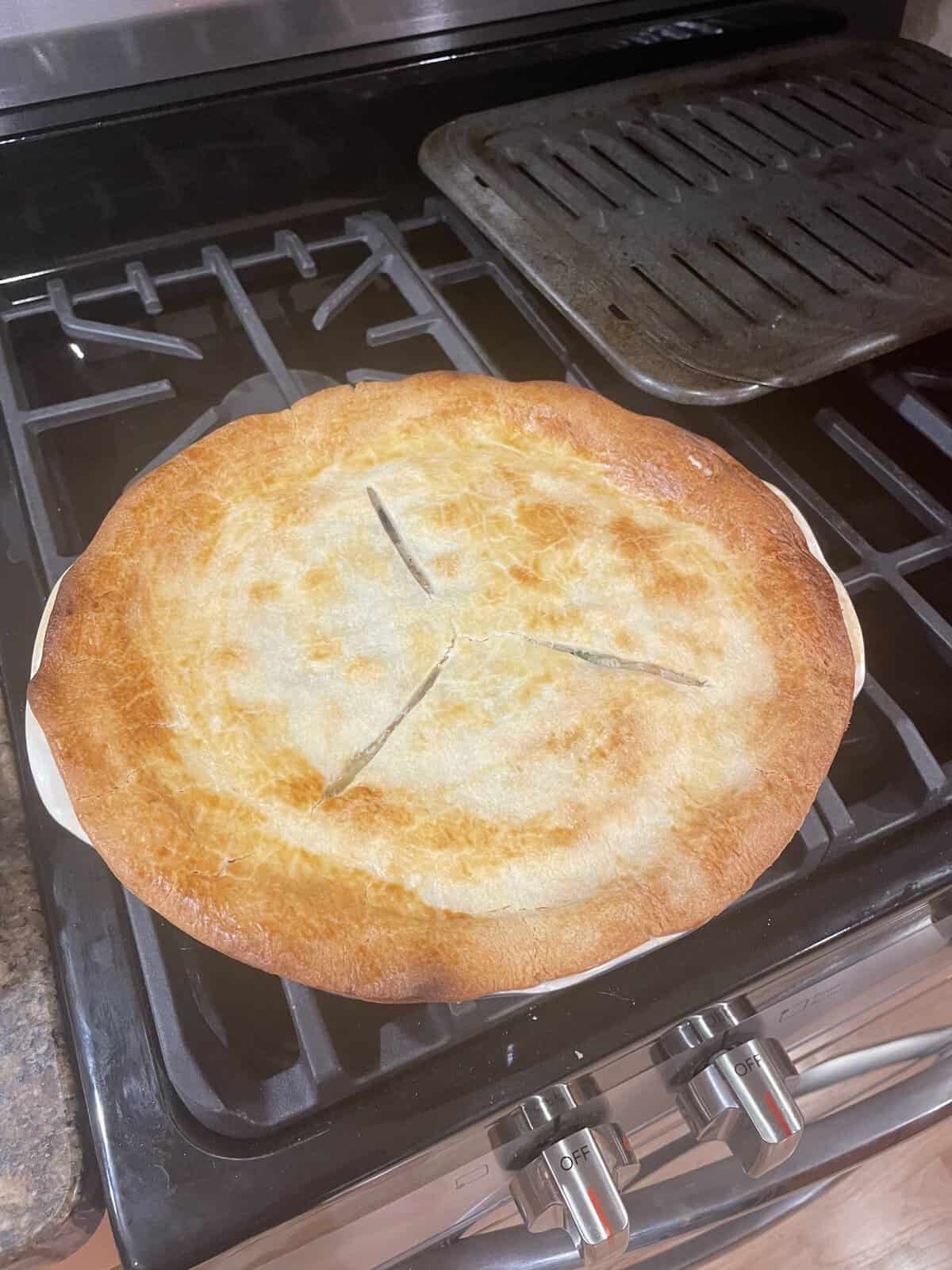 Golden-brown pie with three slits on top, sitting on a stovetop near a metal tray and oven knobs.