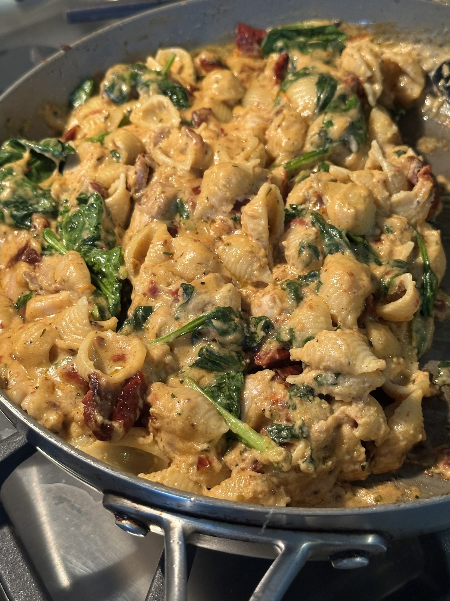 Creamy pasta shells with spinach and sun-dried tomatoes cooking in a skillet on a stovetop.