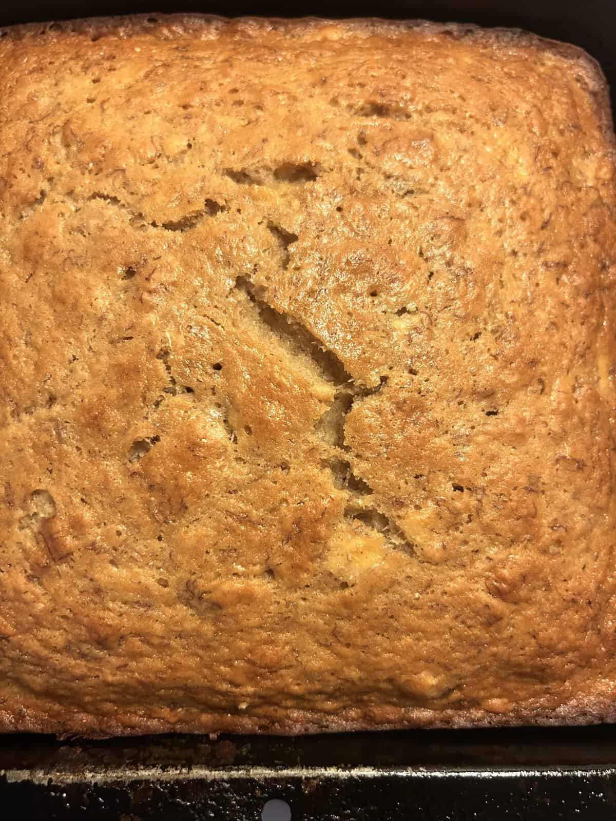 Golden brown banana bread loaf with a large crack running across the top, shown in a baking pan.