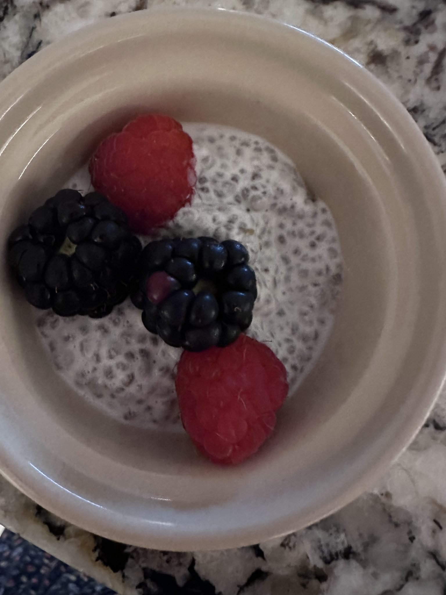 A small bowl of chia pudding topped with two raspberries and one blackberry on a marbled countertop.