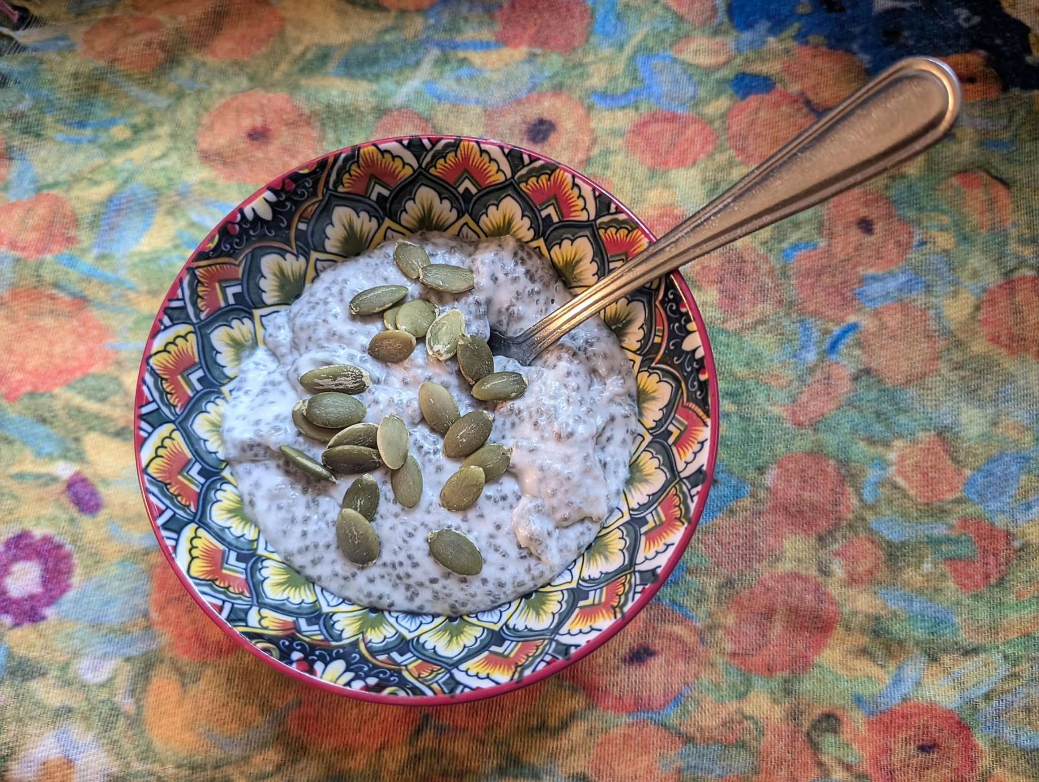 A bowl of chia pudding topped with pumpkin seeds, with a spoon, on a colorful floral tablecloth.