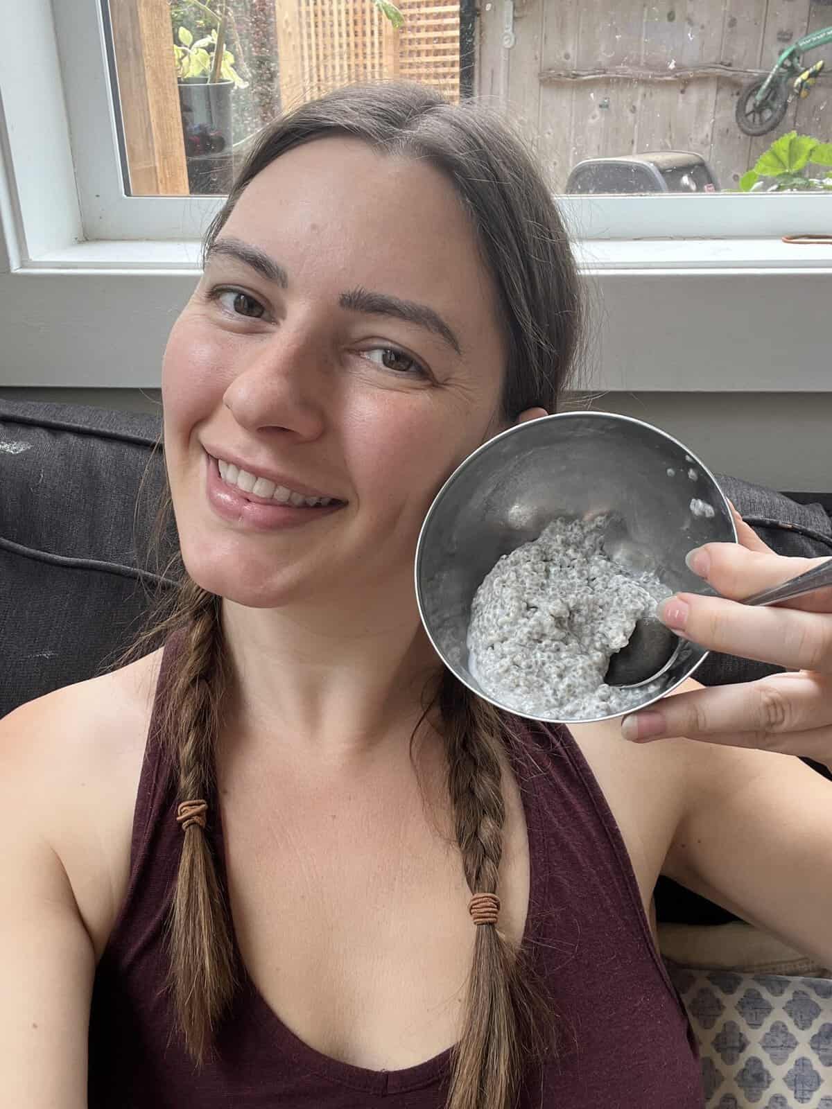 A woman with braided hair smiles while holding a bowl of chia pudding near her face.