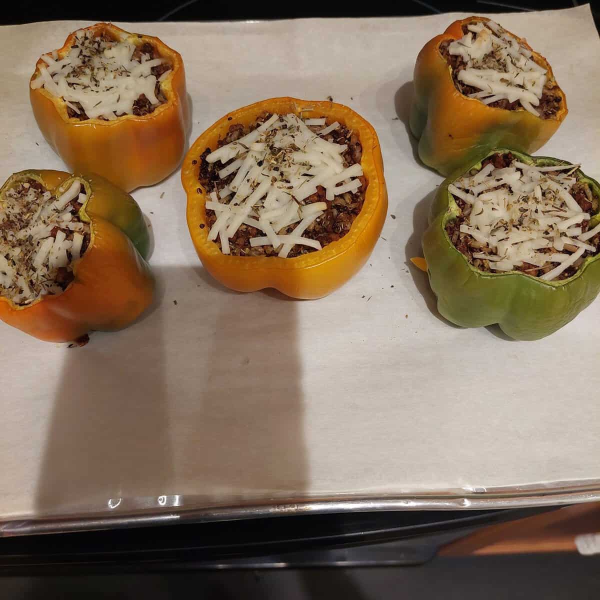Five stuffed bell peppers filled with ground meat and topped with shredded cheese on a baking sheet lined with parchment paper.