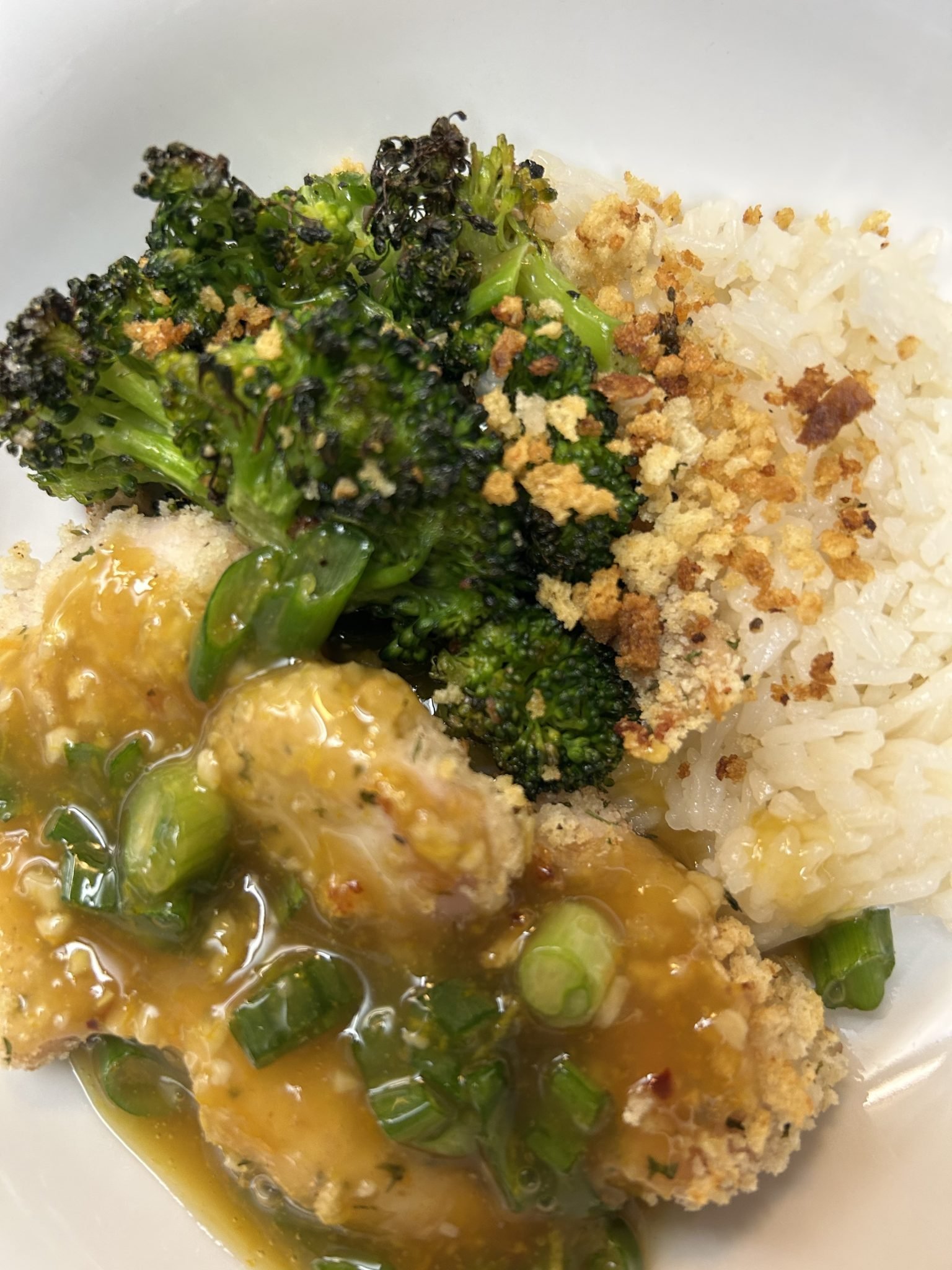 A bowl with white rice, roasted broccoli, bread crumbs, and chicken topped with orange sauce and green onions.