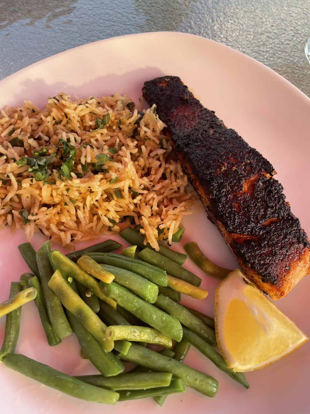 A plate with blackened salmon, green beans, rice, and a lemon wedge on a white dish.