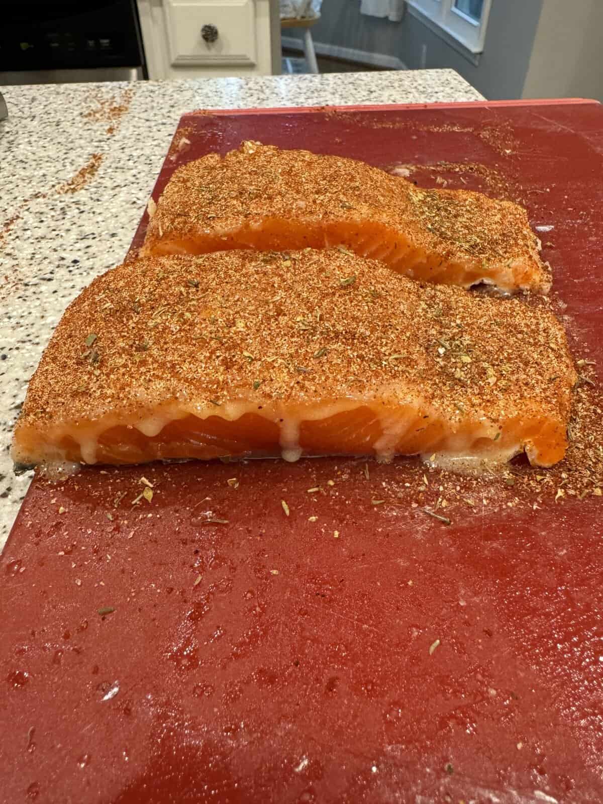 Two raw salmon fillets coated with spices sit on a red cutting board on a kitchen countertop.