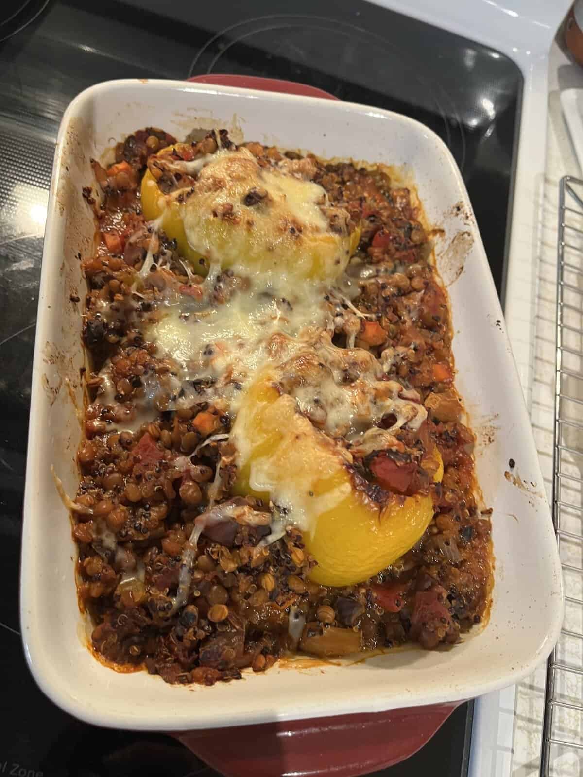 Baked stuffed yellow peppers topped with melted cheese in a white casserole dish with vegetables and lentils.