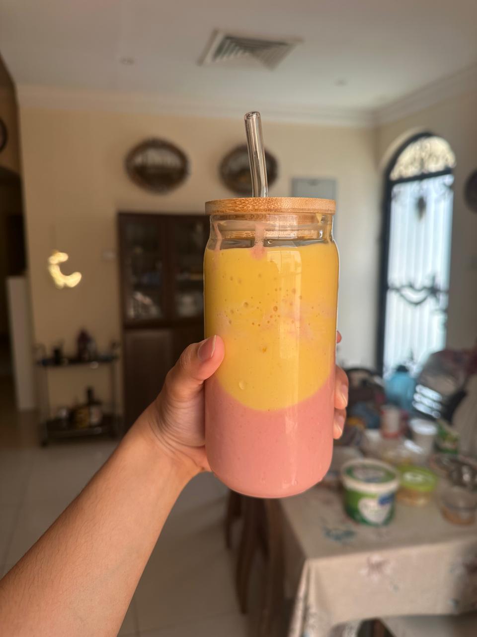 A hand holds a glass jar with a wooden lid and glass straw, filled with a two-layer smoothie: yellow on top and pink on the bottom. The background shows a living room and dining table with some items on it.