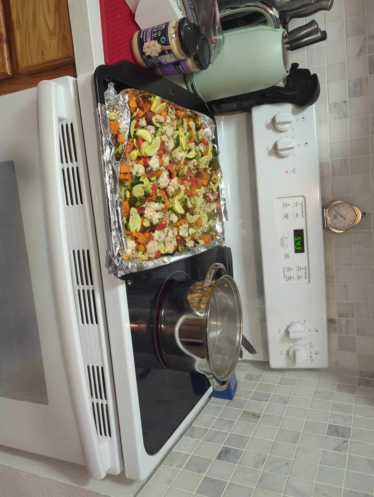 A baking tray of chopped vegetables on foil sits atop a stove, with a pot on a burner and kitchen items nearby.