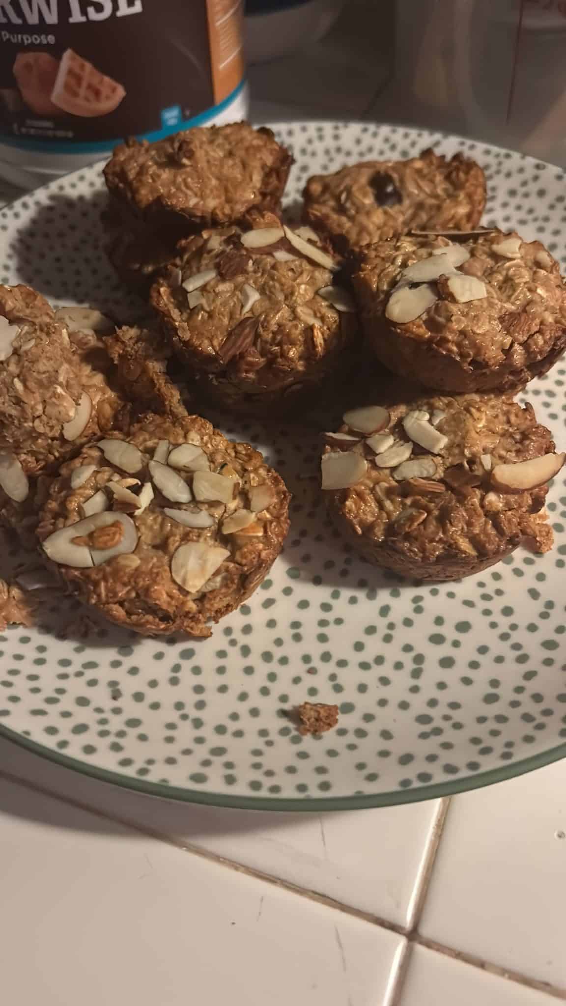 Oat muffins topped with sliced almonds on a white plate with green dots, placed on a kitchen counter.
