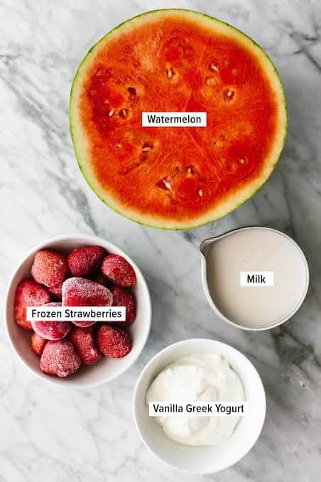 A half watermelon, milk, frozen strawberries, and vanilla Greek yogurt on a marble surface.