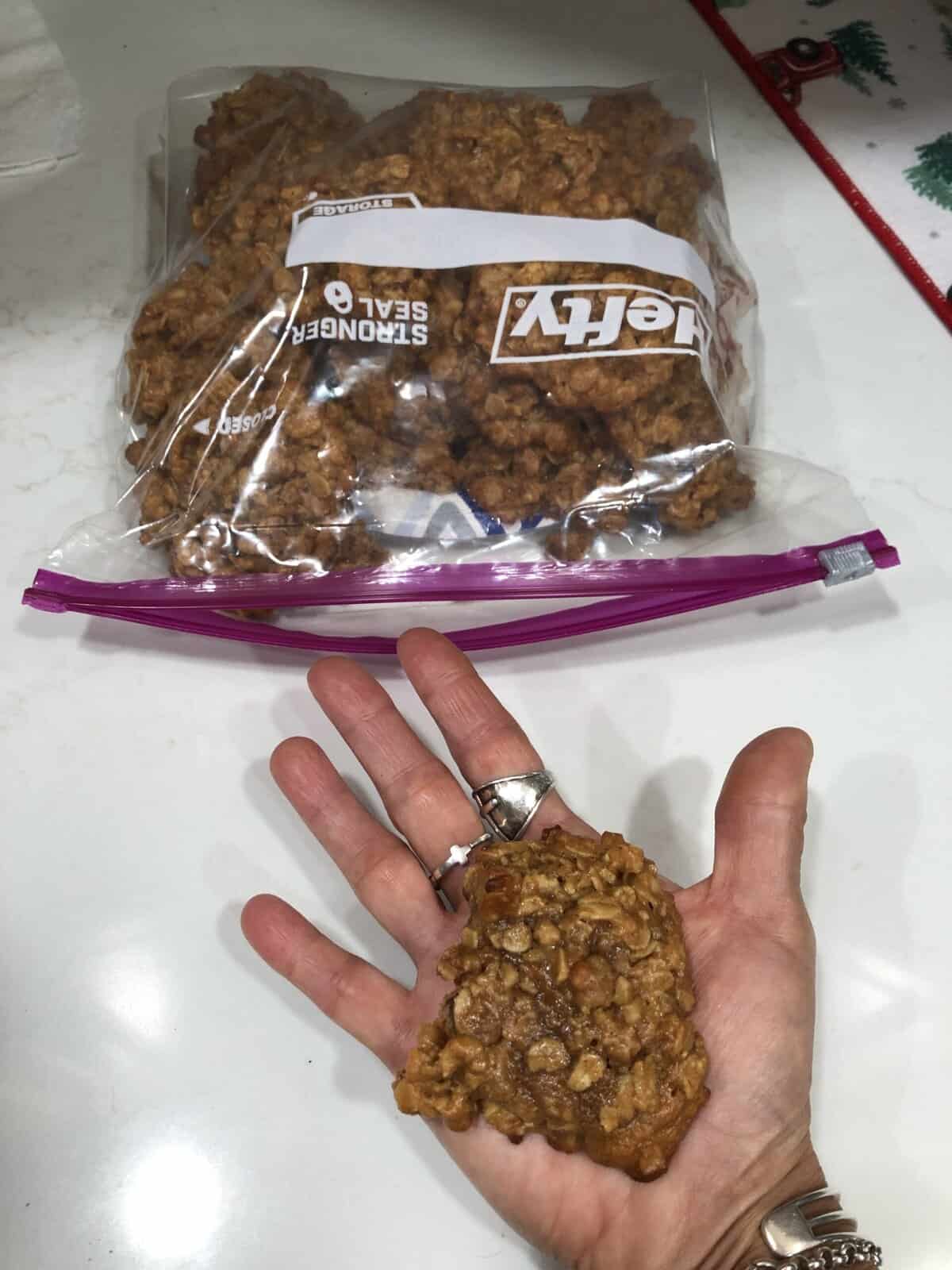 A hand holds a homemade oatmeal cookie above a bag filled with more cookies on a white countertop.