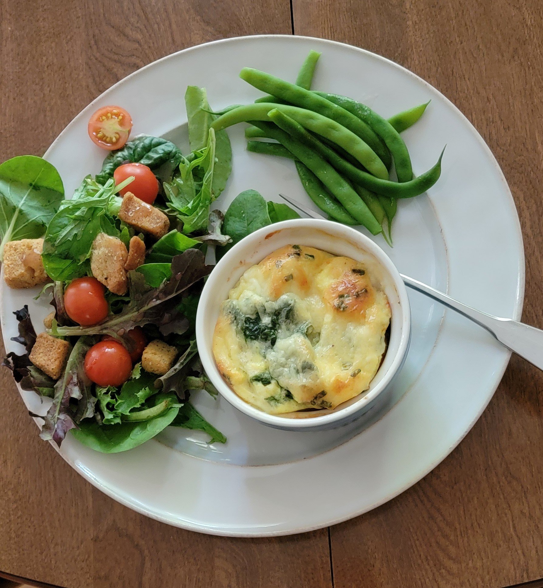 A plate with salad, green beans, and a ramekin of baked egg dish on a wooden table.