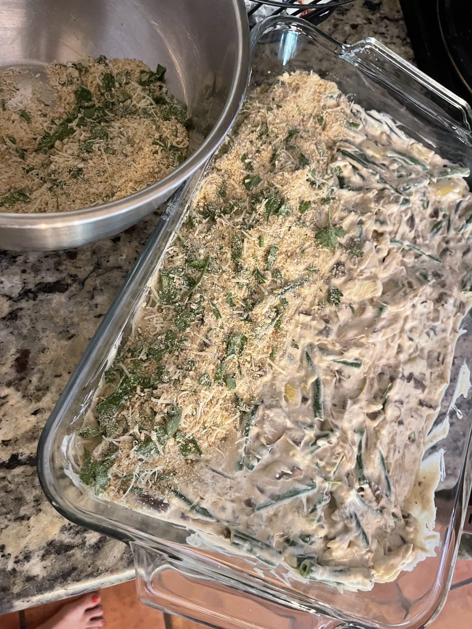 Glass baking dish with creamy green bean casserole, topped with herbs and breadcrumbs, next to a mixing bowl on a counter.