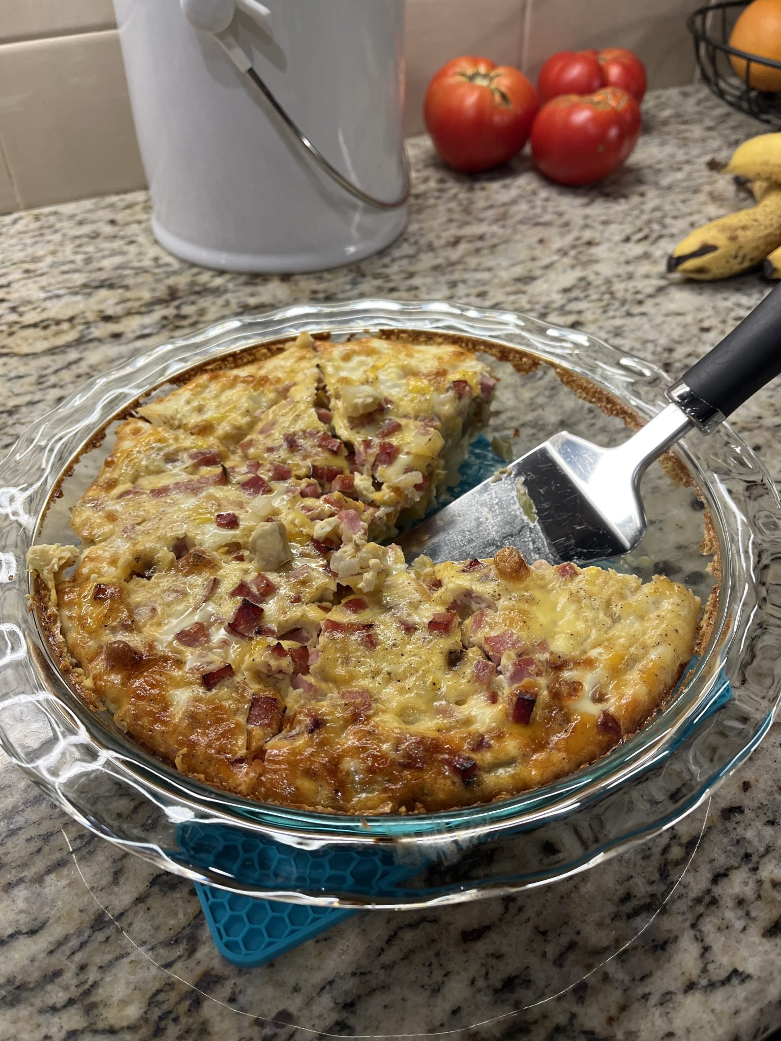 A quiche with ham and cheese in a glass pie dish, one slice missing, on a granite kitchen counter.