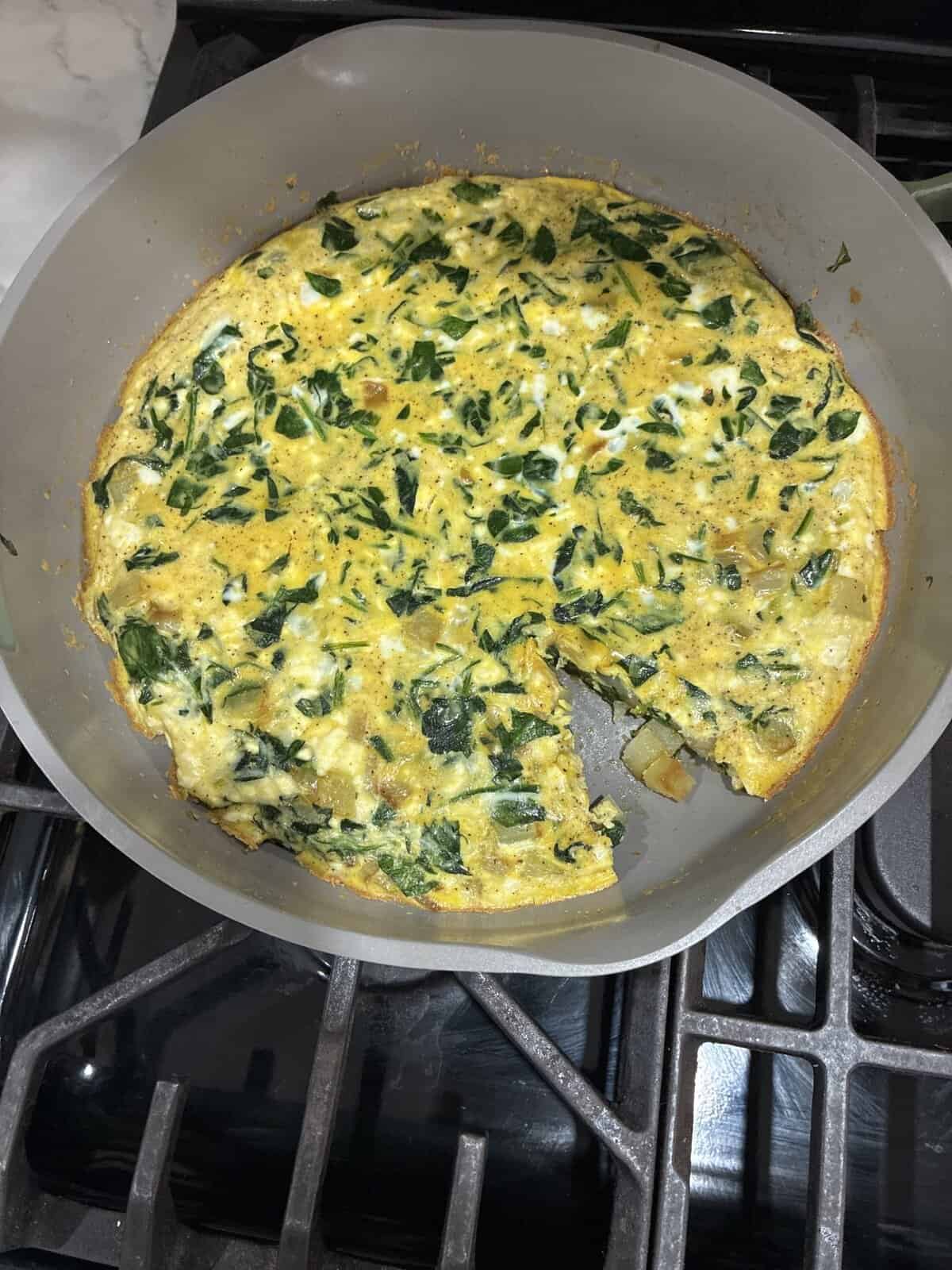 A vegetable frittata in a gray pan with a slice missing, sitting on a stove.