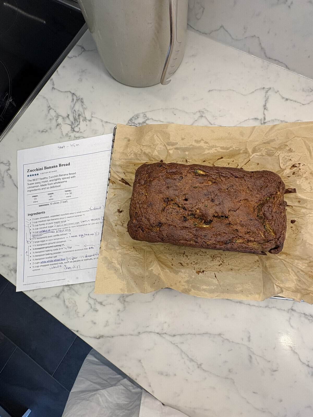 A loaf of zucchini banana bread on parchment paper next to a printed recipe on a white marble countertop.