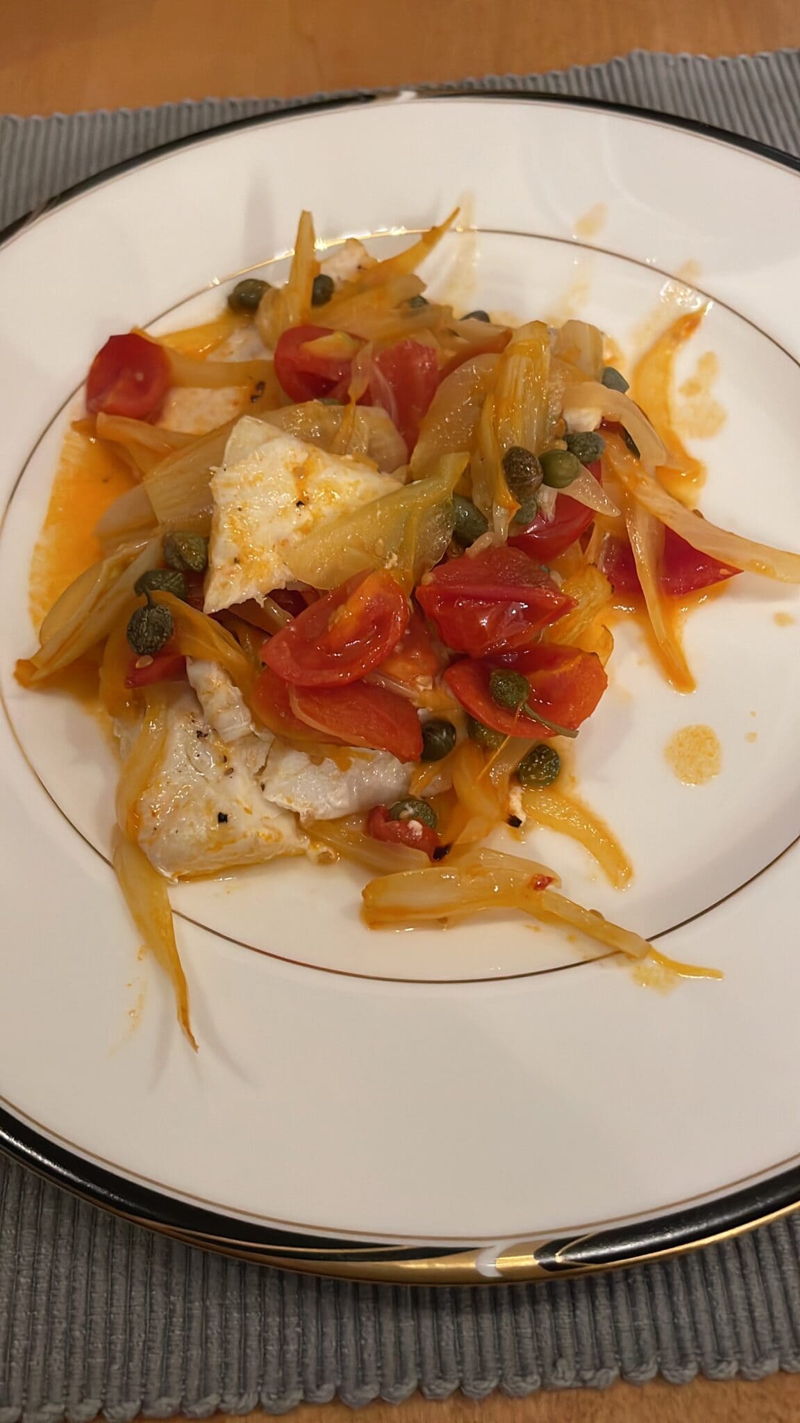 White fish topped with cherry tomatoes, capers, and sliced onions on a white plate with a silver rim.