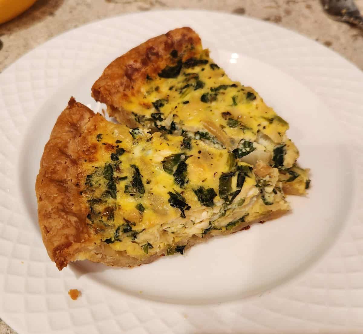 Two slices of spinach quiche with a golden crust on a white plate, sitting on a granite countertop.