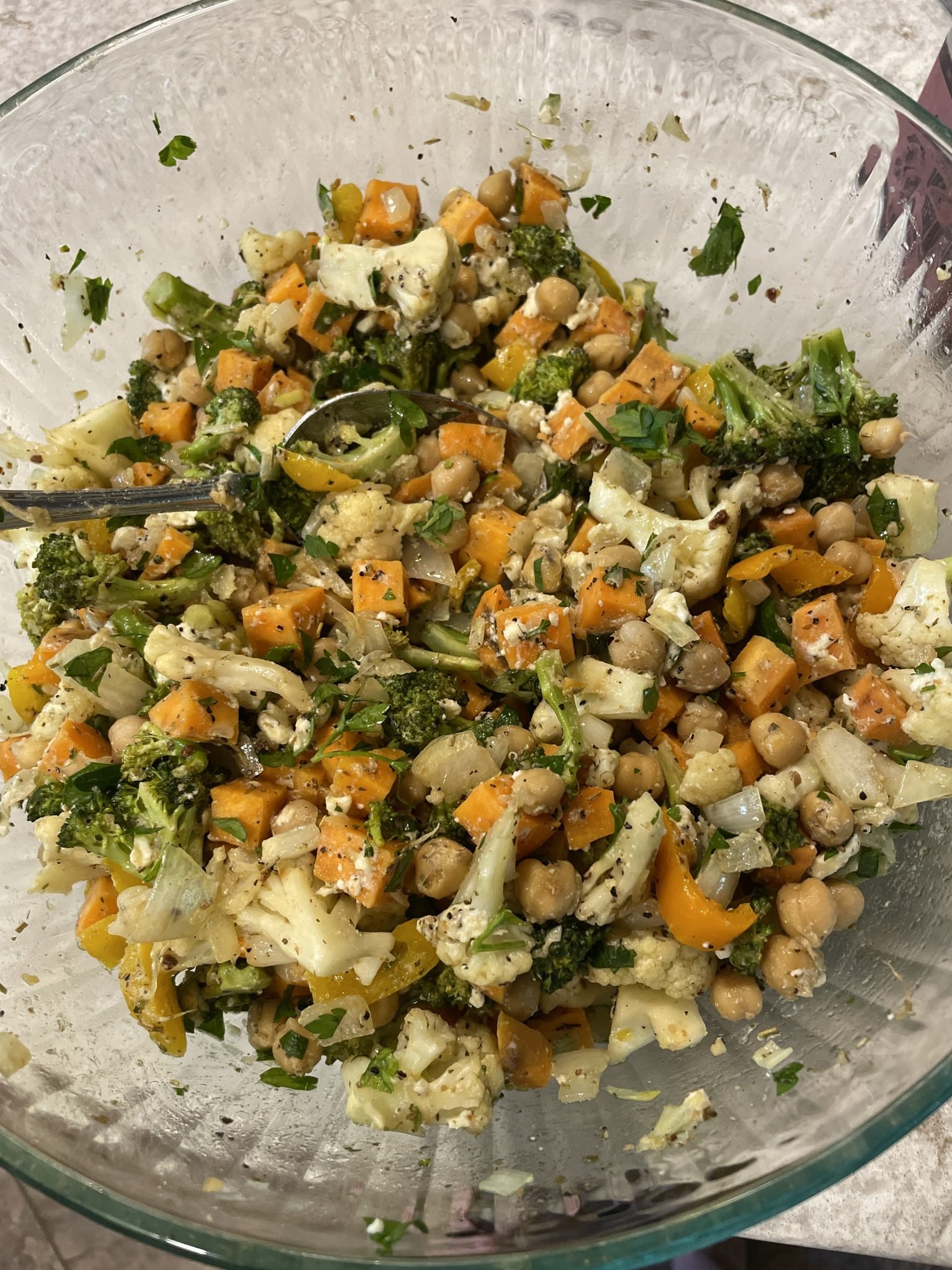 A glass bowl filled with a colorful salad made of chickpeas, chopped broccoli, cauliflower, carrots, yellow bell peppers, and fresh herbs. A spoon is in the bowl, ready for serving.