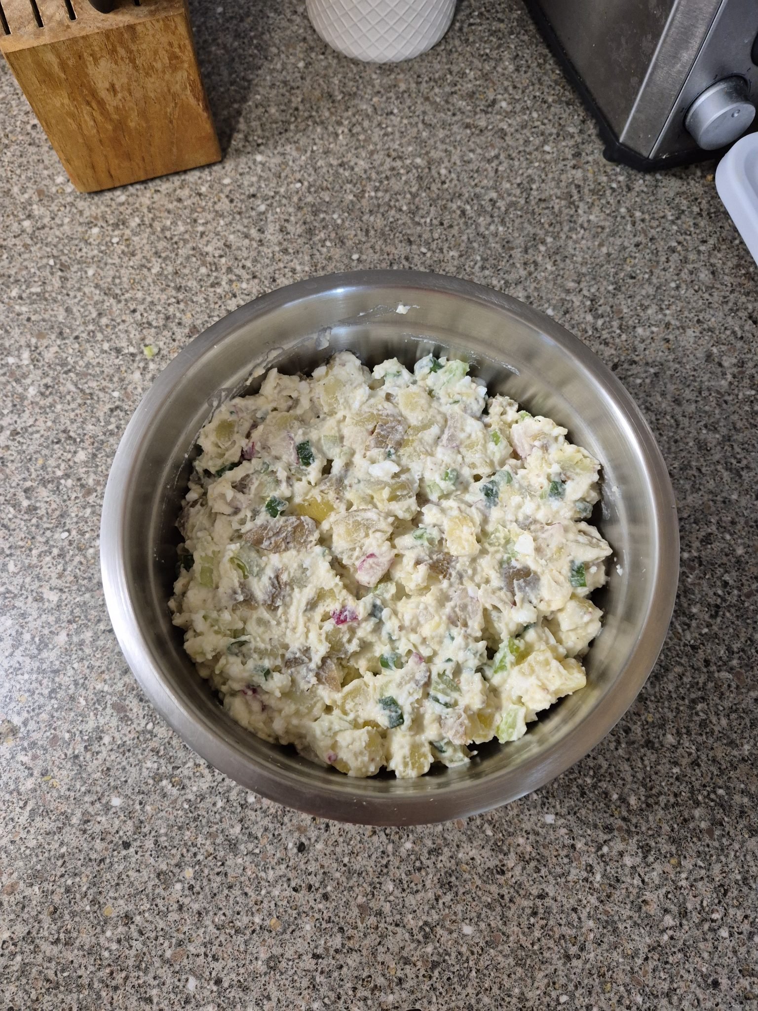 A stainless steel bowl of potato salad with herbs and creamy dressing sits on a kitchen countertop.