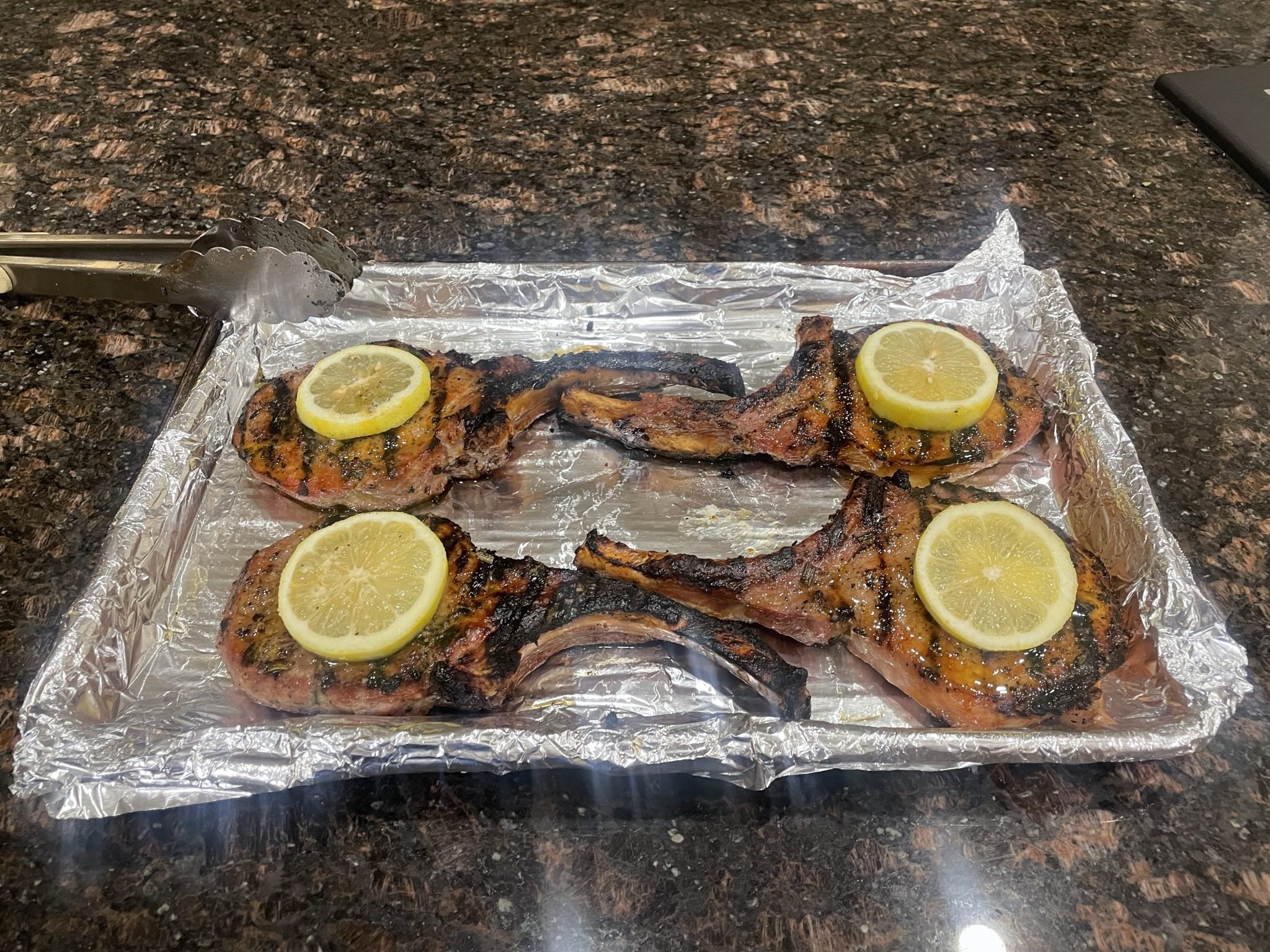 Four grilled lamb chops with lemon slices on top, placed on a foil-lined baking tray on a granite counter.