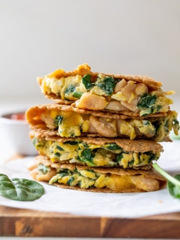 Stacked breakfast quesadillas filled with spinach, scrambled eggs, and beans on a wooden board, with a bowl in the background.
