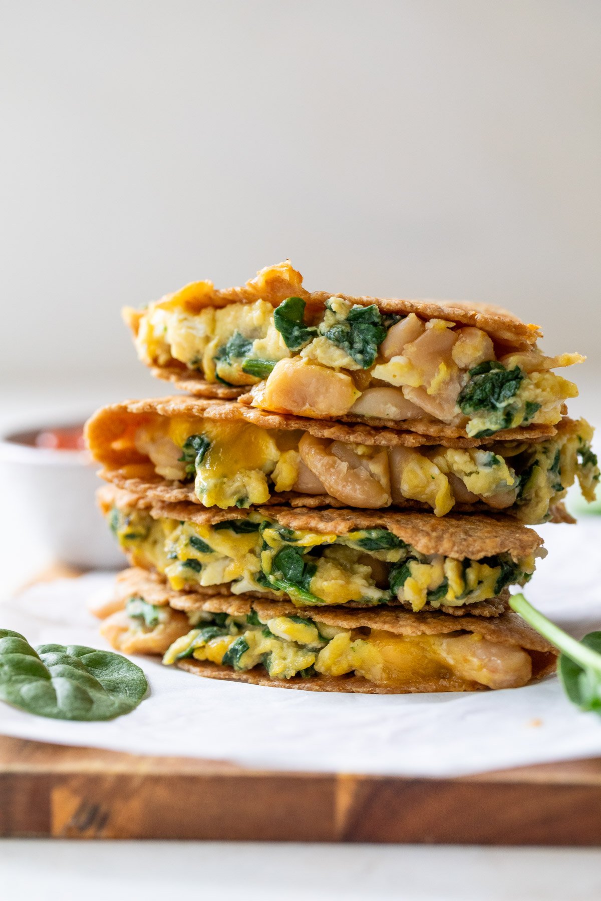 Stacked breakfast quesadillas filled with spinach, scrambled eggs, and beans on a wooden board, with a bowl in the background.