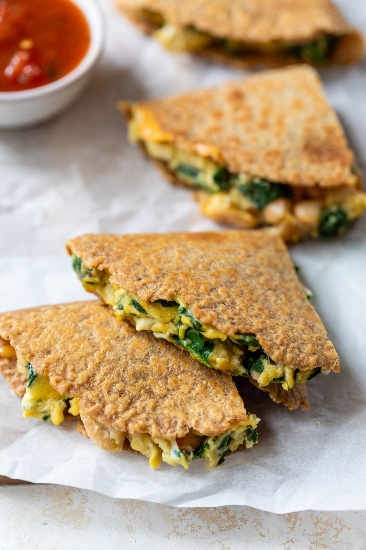 Two golden-brown breakfast quesadilla wedges with spinach and cheese filling on parchment, with salsa in a small bowl nearby.