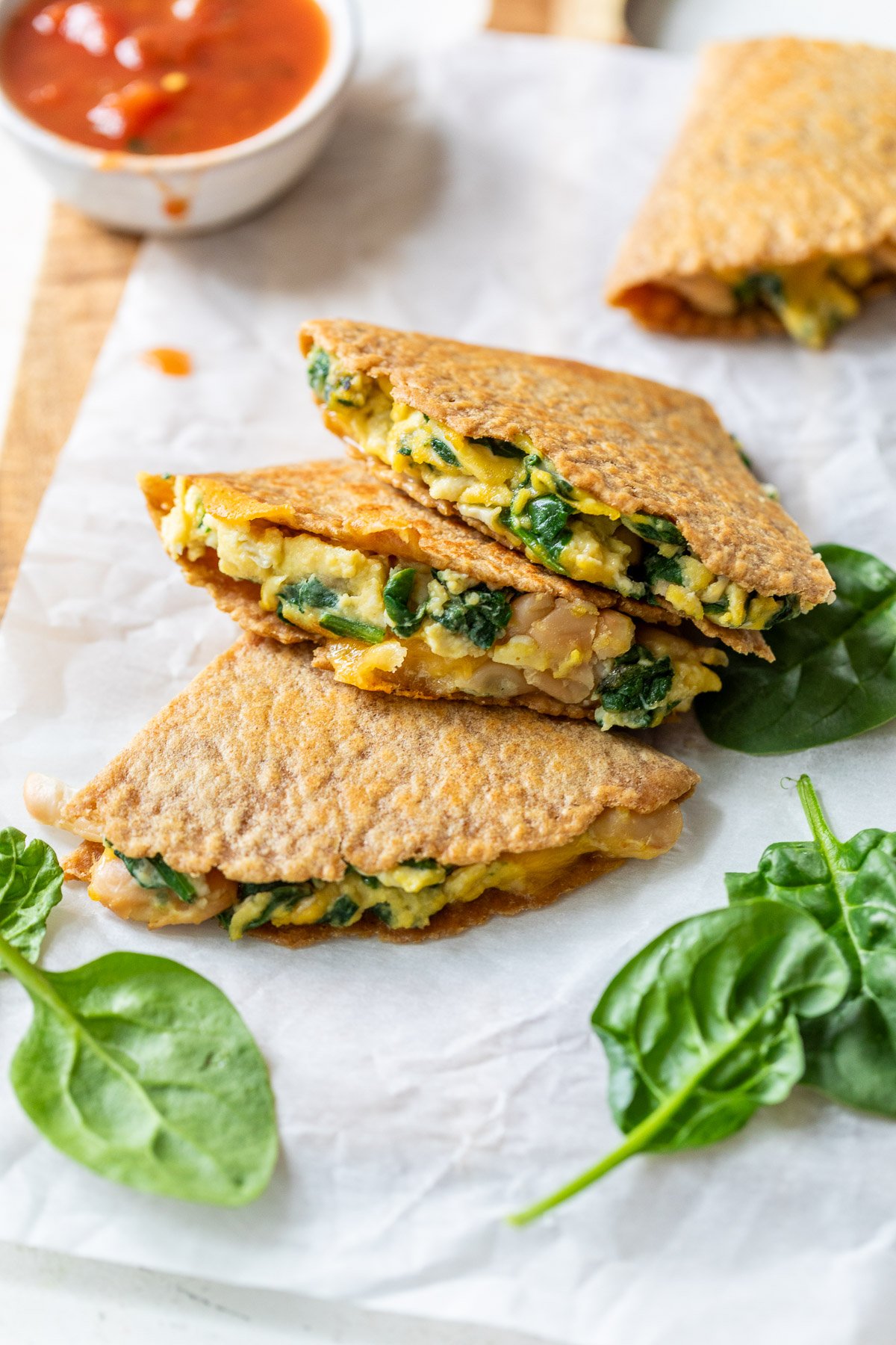 Three folded breakfast quesadillas filled with eggs and spinach on parchment paper, with salsa and spinach leaves nearby.