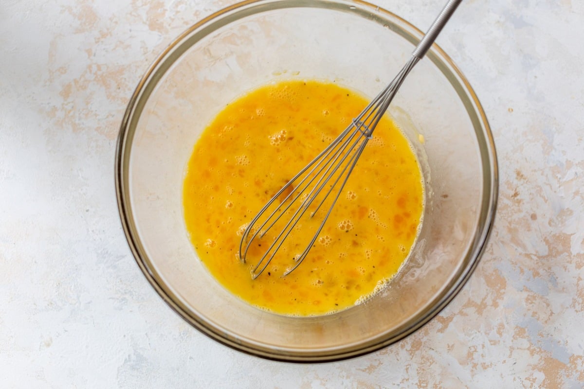 A metal whisk in a glass bowl with beaten eggs on a light, textured surface.