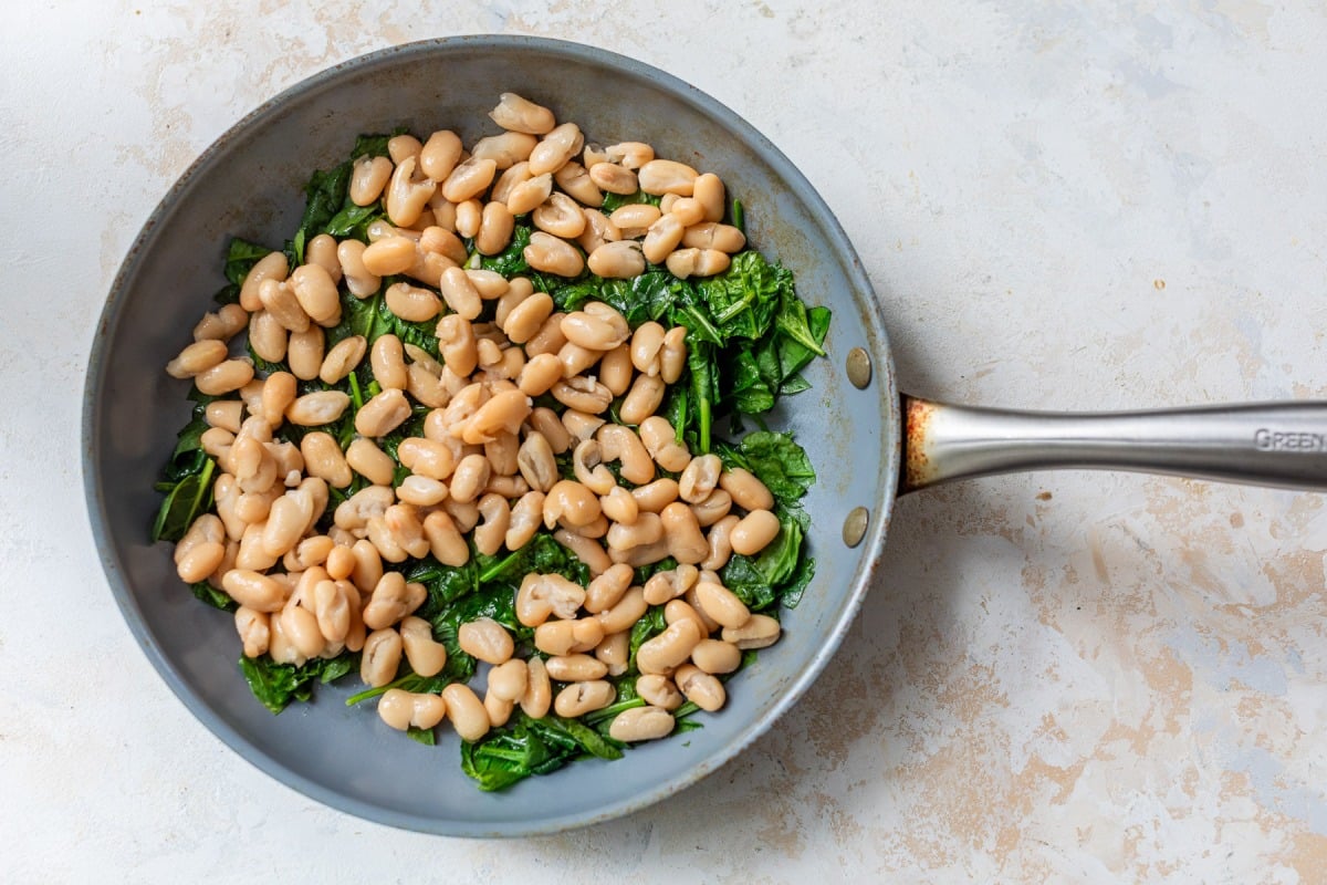 White beans and sautéed greens in a silver skillet on a light-colored surface.