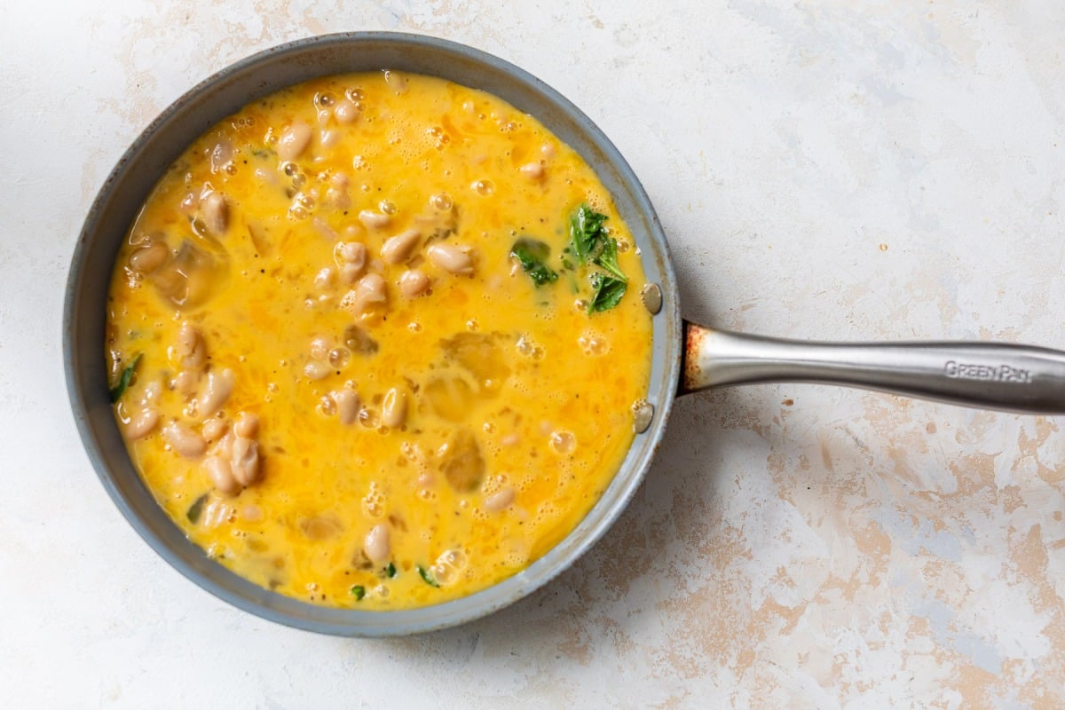 A frying pan with beaten eggs, white beans, and greens, ready to be cooked on a light countertop.