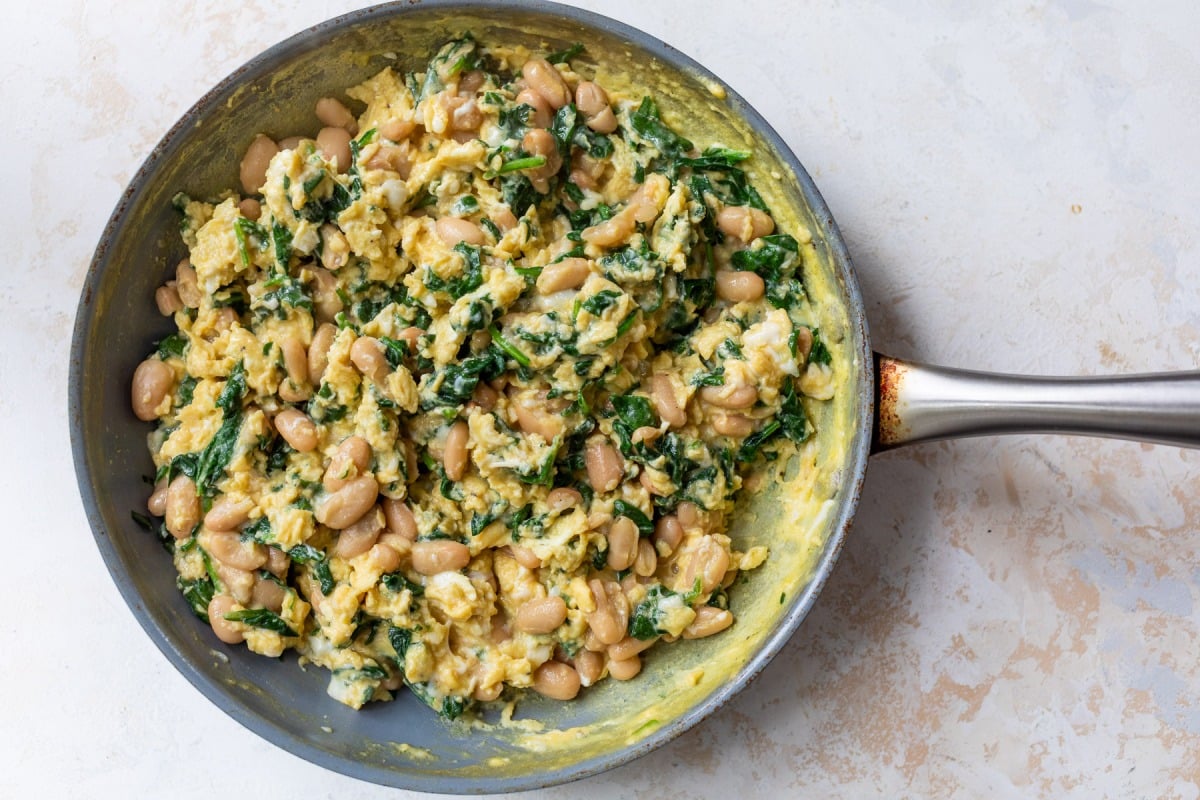 Scrambled eggs with white beans and spinach in a frying pan on a light background.