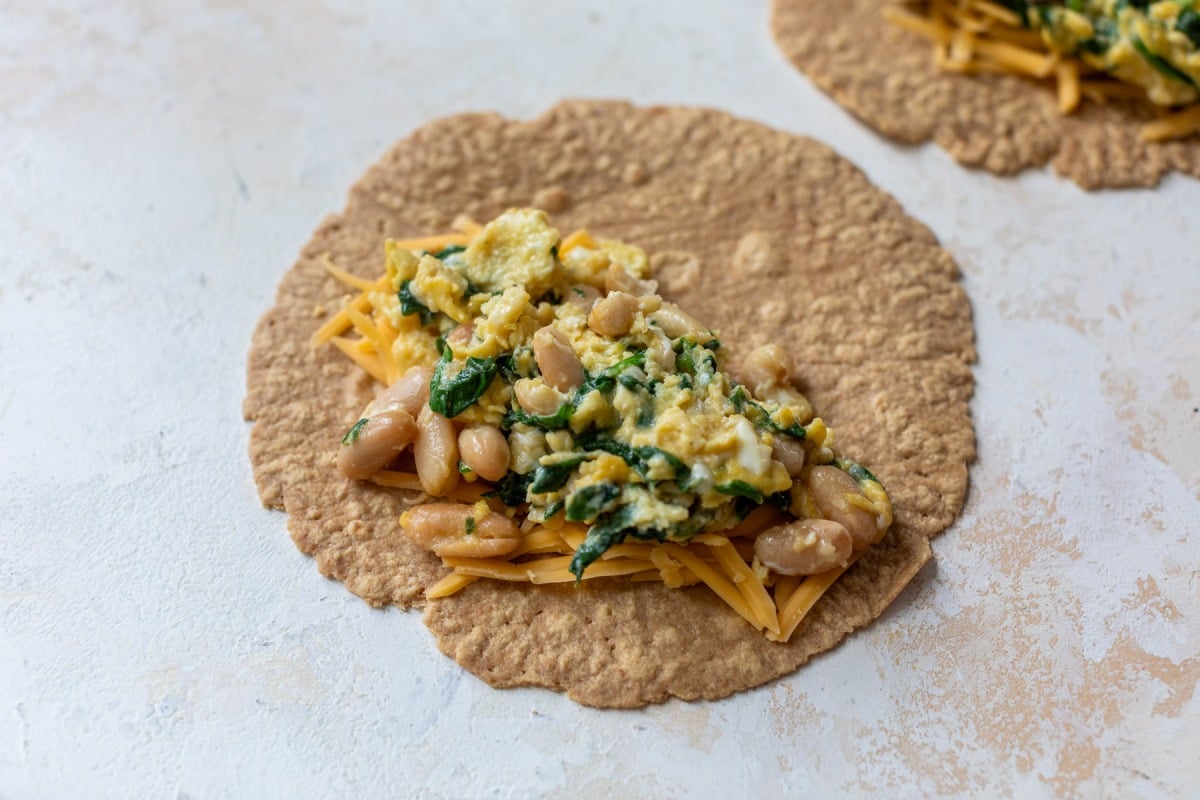 A whole wheat tortilla topped with scrambled eggs, beans, spinach, and shredded cheddar cheese on a light surface.