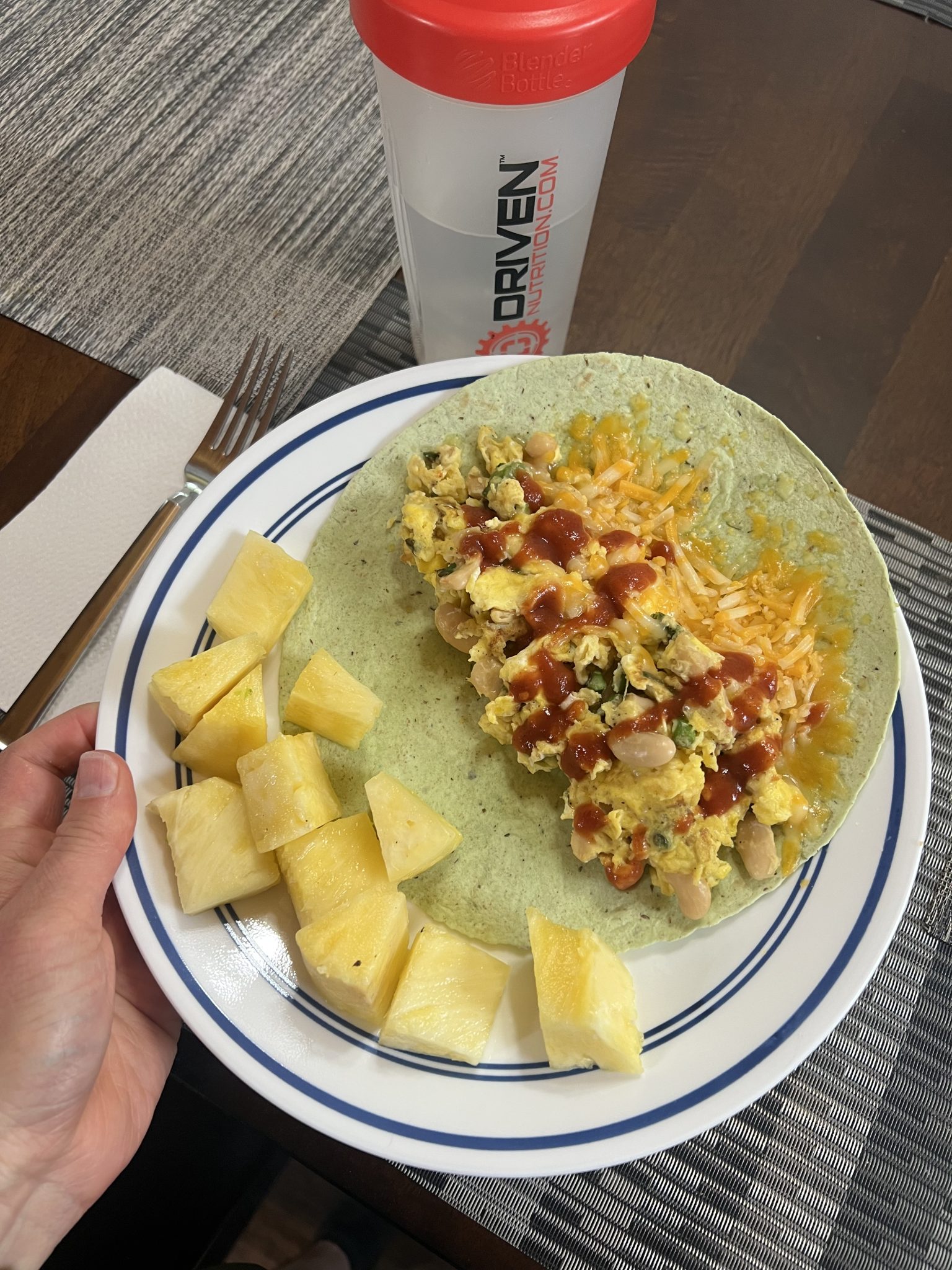 A hand holds a plate with a spinach tortilla topped with scrambled eggs, beans, cheese, and salsa, alongside pineapple chunks. A red and white shaker bottle and a napkin are on the table.