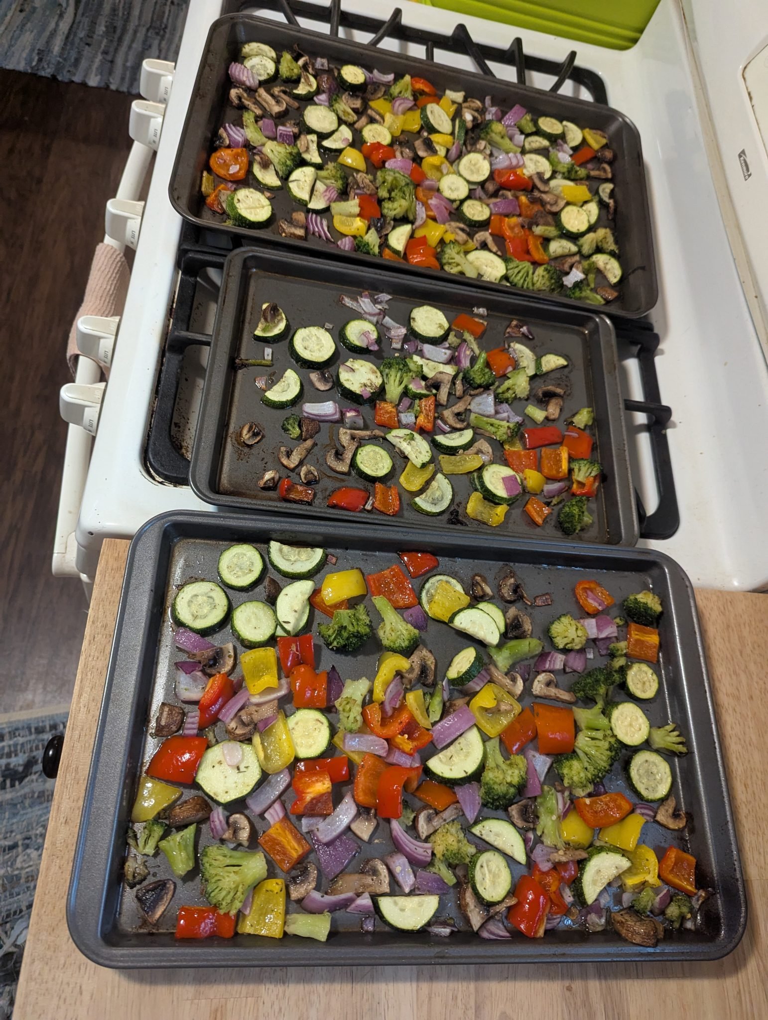 Three baking sheets with chopped vegetables, including zucchini, broccoli, peppers, and onions, ready to be roasted.