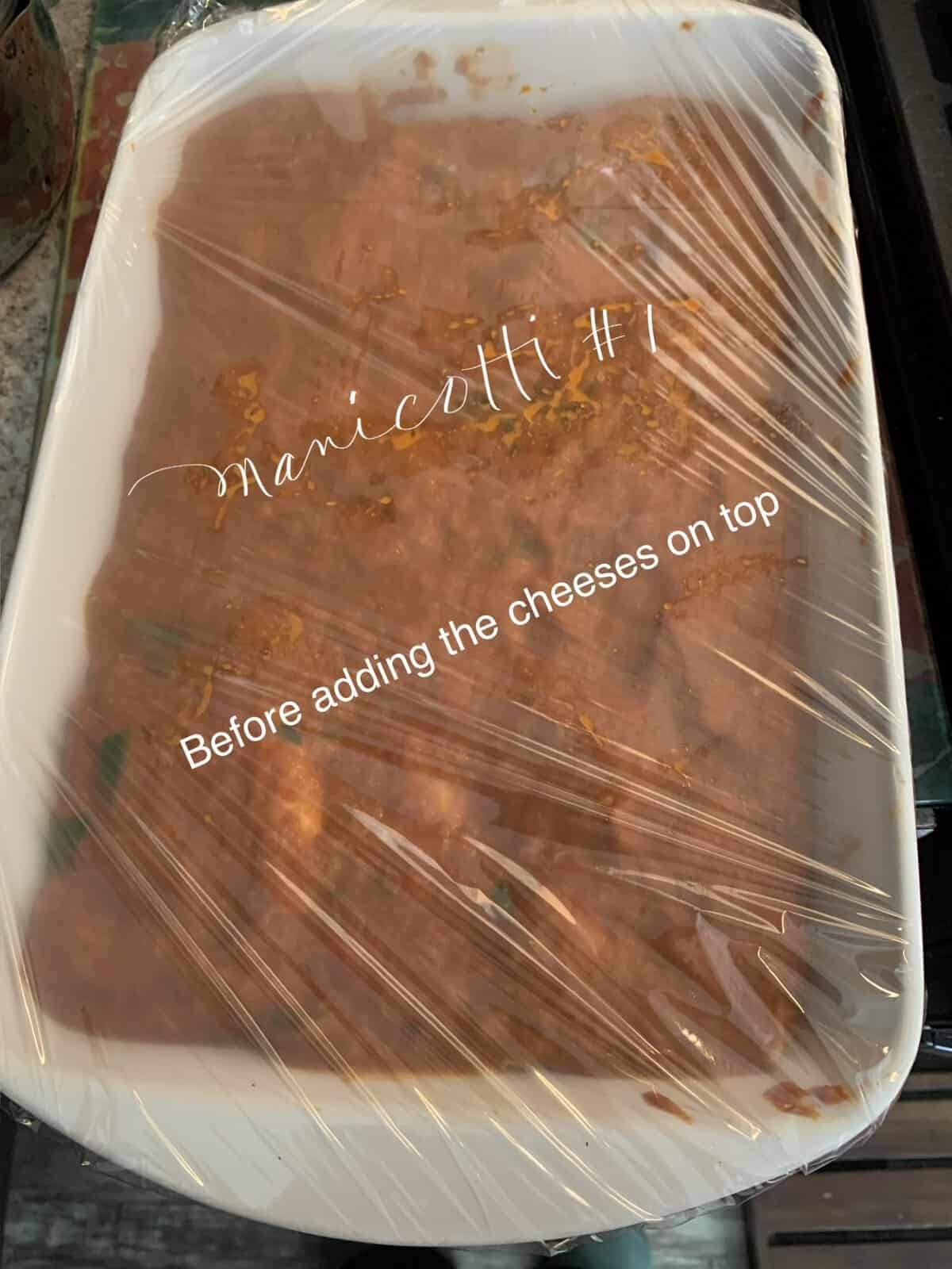 A casserole dish covered in plastic wrap, filled with manicotti in sauce, labeled "Before adding the cheeses on top.