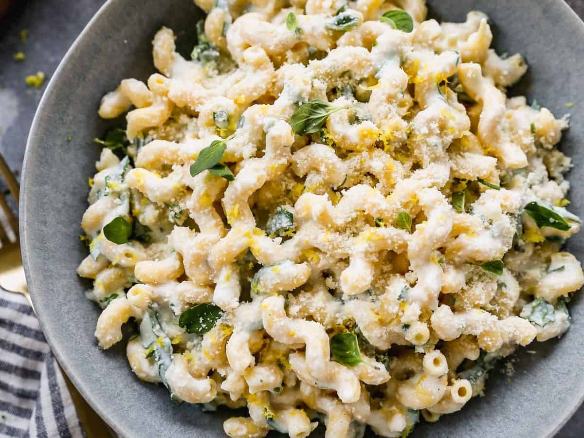 A bowl of creamy pasta with a sprinkle of herbs and grated cheese. The pasta is set on a gray surface with a striped cloth, golden fork, and some greens nearby. The dish looks rich and garnished with fresh herbs and lemon zest.