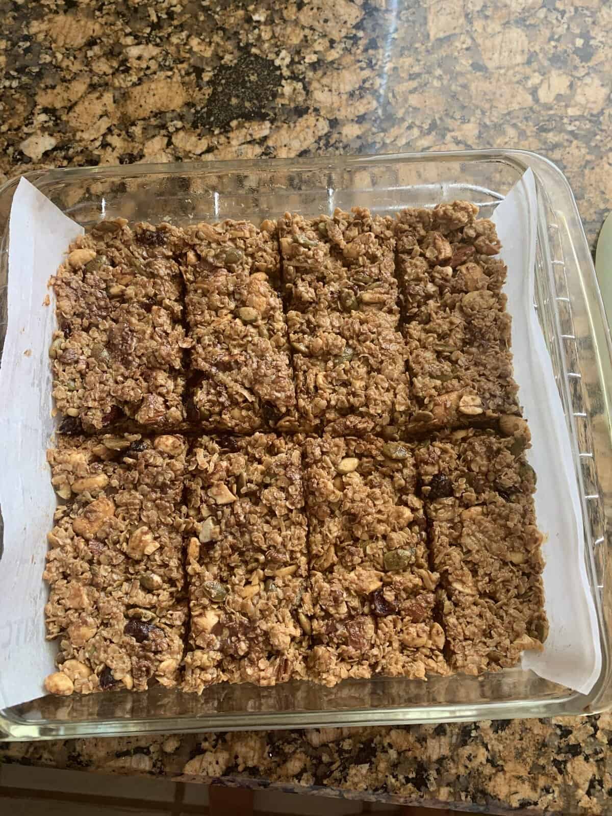 A glass pan with parchment paper holds twelve homemade granola bars cut into squares on a granite countertop.