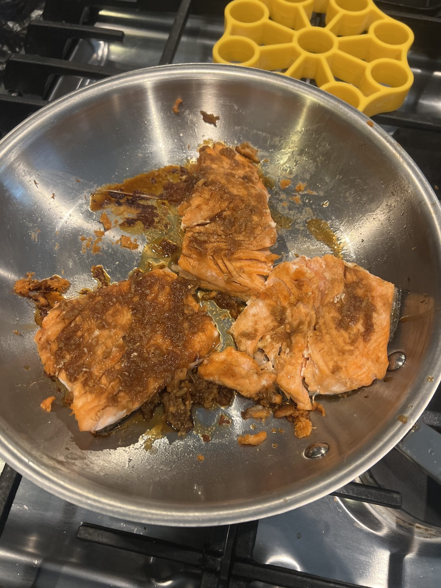 Cooked salmon fillets with browned seasoning in a stainless steel pan on a stovetop, yellow mold nearby.