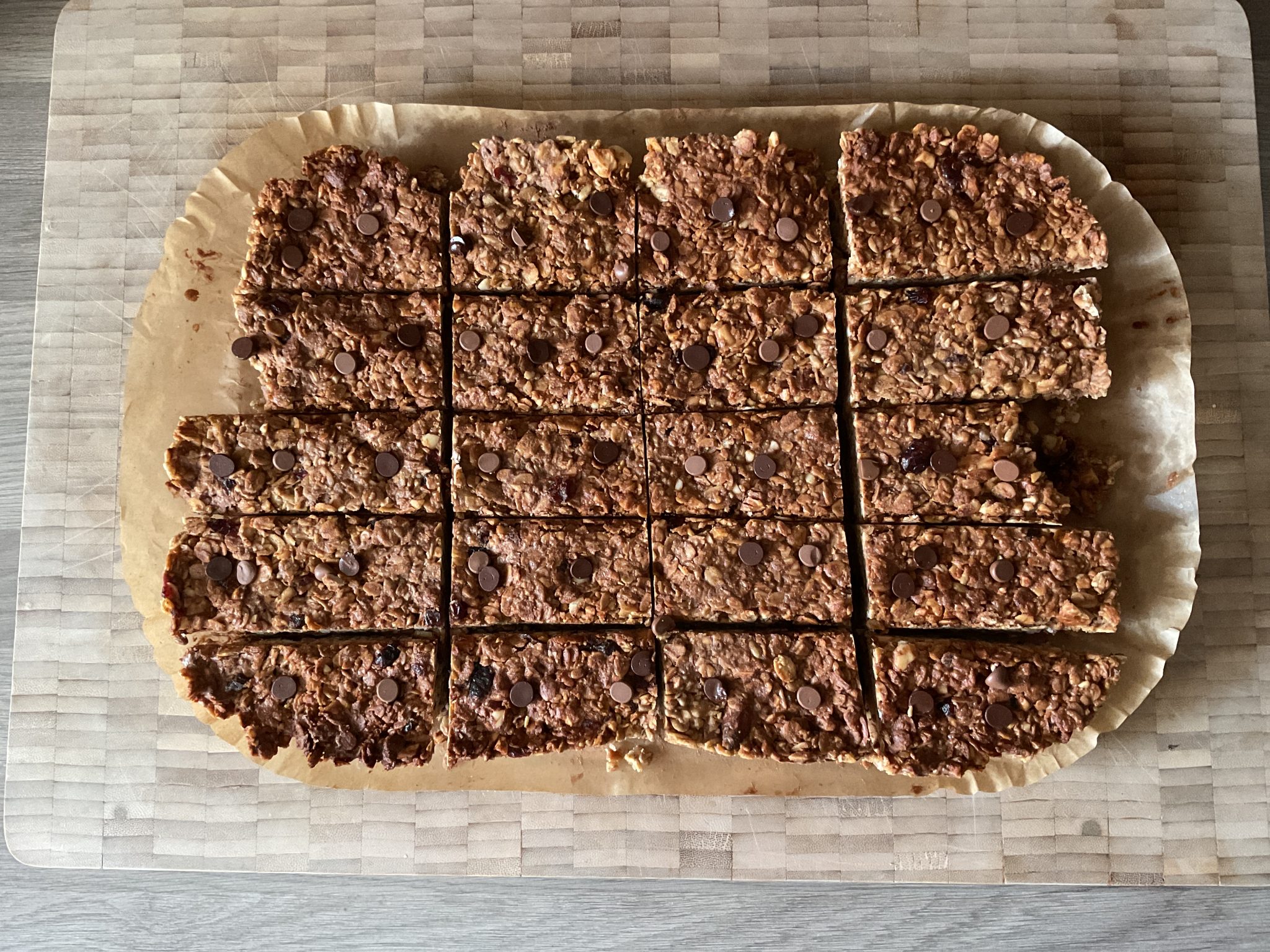 Sixteen chocolate chip oat bars arranged on parchment paper on a wooden cutting board.