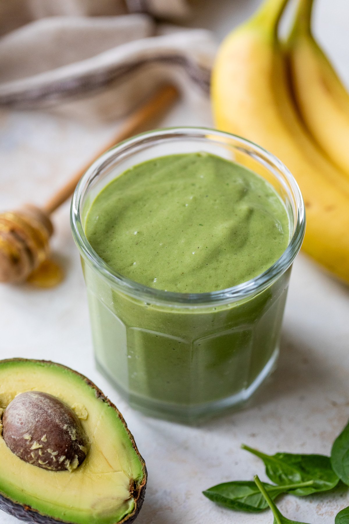 A glass filled with green avocado smoothie, surrounded by a halved avocado, fresh spinach leaves, a banana, and a honey dipper with honey.